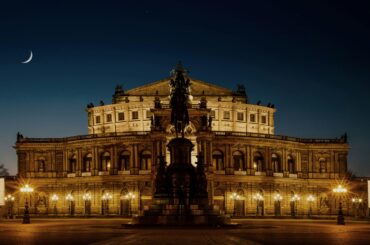 Beautifully illuminated Semper Opera House in Dresden at night with a crescent moon in the sky.