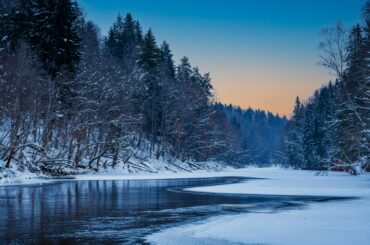 A tranquil winter scene of a snow-covered river in Cēsis, Latvia at dusk.
