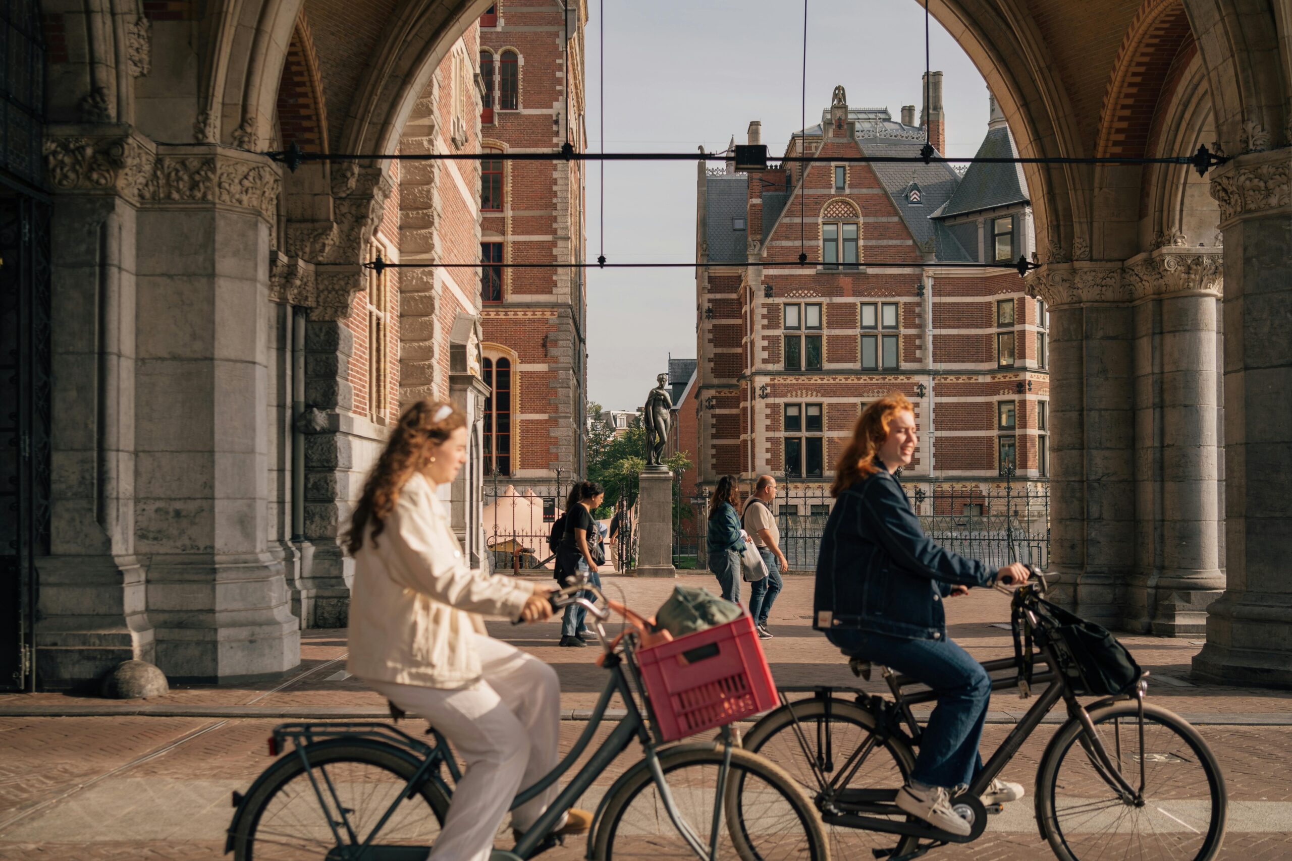 Women cycling under an Amsterdam archway with historical buildings in the background.