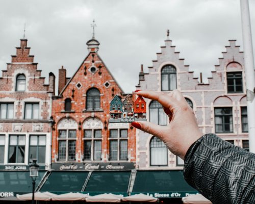 A hand holding a miniature Bruges building with traditional Belgian architecture in the background.