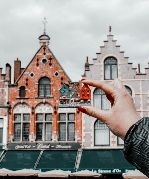 A hand holding a miniature Bruges building with traditional Belgian architecture in the background.
