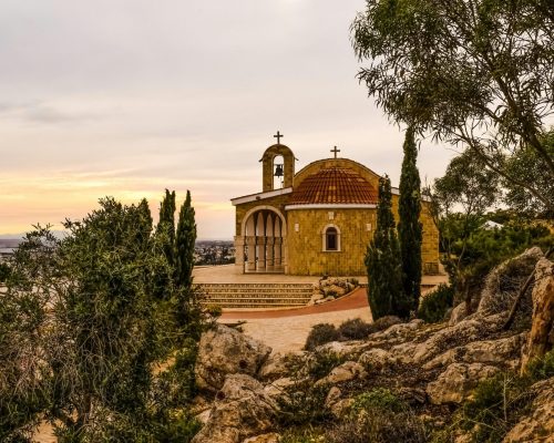 A picturesque historic church in Cyprus at sunset, surrounded by trees and rocks.