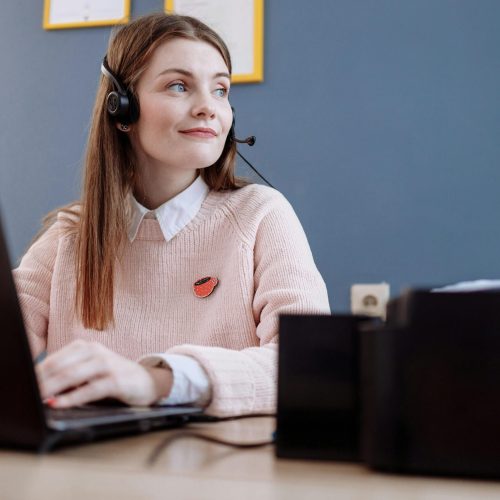 A cheerful woman in a pink sweater working remotely with a laptop and headset, providing customer service.