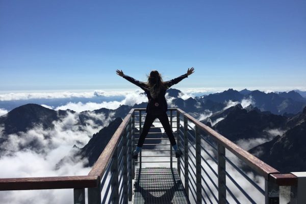 A woman stands on a viewing platform in Vysoké Tatry, Slovakia, surrounded by clouds and mountains, embracing freedom.