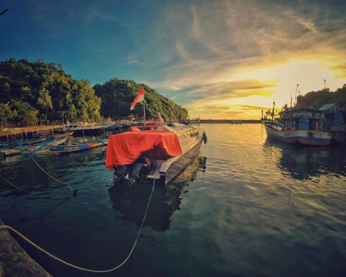 A stunning sunset view at Girisubo Harbor, Indonesia, with boats and lush hills.
